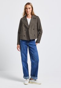 Avklippt, dubbelknapad jacka i olivgrön tyg med svarta knappar. Matchad med blå jeans och vita sneakers. Enkel, ren design.