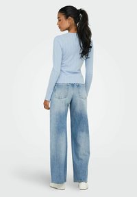 Modell, das von der Kamera weg schaut, trägt einen hellblauen Rippenpullover und weit geschnittene, ausgebleichte Jeans, vor einem schlichten weißen Hintergrund.