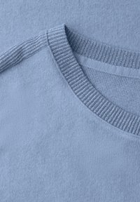 Světlounce modrá pletená látka s hladkou texturou. Má kulatý výstřih s žebrovaným detailem. Detailní záběr na šití.