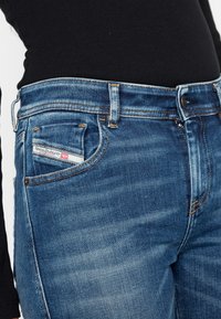 Jeans de mezclilla en azul medio con una textura desgastada, que cuentan con un bolsillo para monedas, costuras en color naranja y una etiqueta de marca en el bolsillo trasero.