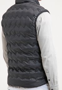 Chaleco acolchado negro con un patrón de rombos, cuello alto y mangas. El material parece suave y resistente al agua.