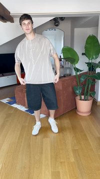 Camiseta beige de gran tamaño con un acabado brillante, combinada con pantalones cortos de denim negros holgados. Zapatillas blancas que complementan el atuendo casual. Entorno interior con plantas.