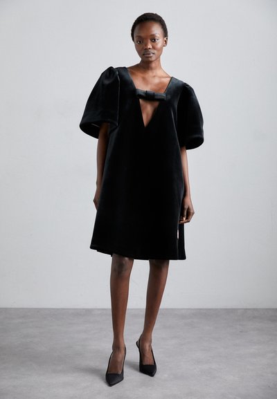 Black velvet dress with a deep V-neck and oversized puffy sleeves, featuring a bow accent at the neckline. Worn with black pointed heels.