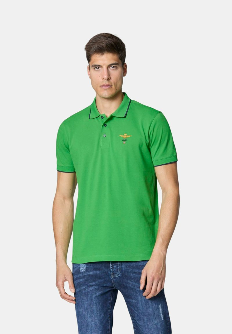 Camisa polo verde con cuello, que presenta un ribete en contraste en color azul marino, un logo bordado en el pecho y mangas cortas. Se lleva con unos pantalones vaqueros azules.