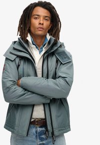 Jasnoniebieska kurtka ocieplana z kieszeniami na zamek, wysokim kołnierzem i haftowanym logo na ramieniu. Nosi ją na szarym swetrze i dżinsach.