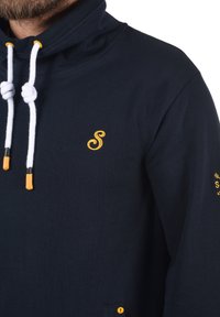 Sudadera de color azul marino con cuello alto, cordones blancos, detalles amarillos y un logo bordado en el pecho. Presenta un bolsillo lateral.
