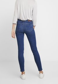 Jeans ajustados de denim azul oscuro con una textura suave, que presentan una cintura de media altura y bolsillos traseros tradicionales. Combinados con zapatillas blancas.