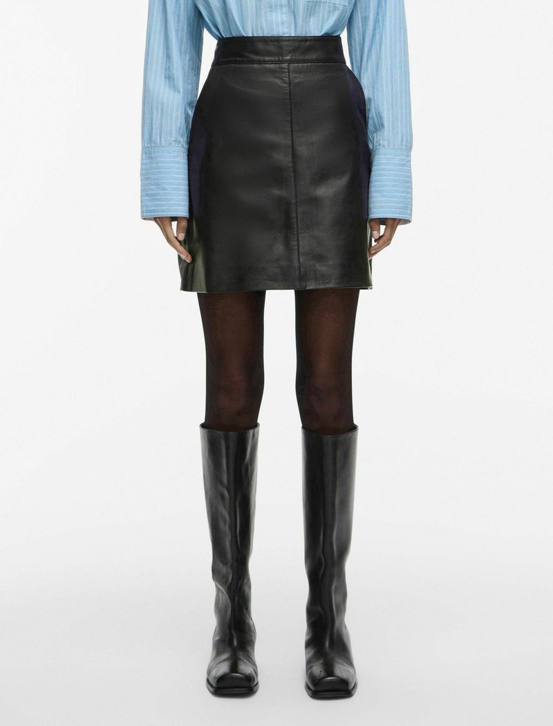 Svart miniskjørt i skinn med høy midje, kombinert med knelang svarte støvler. Skjørtet har en glatt tekstur og tettsittende design.