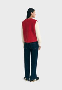 Rode fleece vest met een ronde hals, gedragen over een crèmekleurig shirt met lange mouwen. Gecombineerd met donkerblauwe wijde jeans en zwarte schoenen.