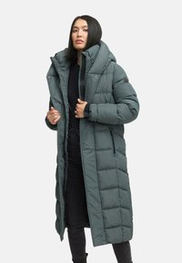 Long, padded coat in muted green with a high collar, zip closures, and side pockets. Features a quilted pattern and soft texture.