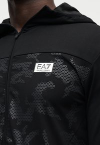 Μαύρο μπουφάν με φερμουάρ, με διακριτικό υφασμάτινο μοτίβο, εφαρμοστή γραμμή και έντονο λογότυπο EA7 στο στήθος.