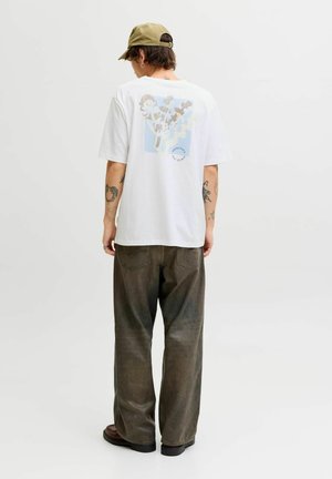 Personne portant un T-shirt blanc à motifs, un pantalon ample foncé, des chaussures marron et une casquette verte, debout de dos face à la caméra sur un fond uni.