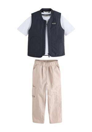 Dreng stående iført en sort vest over en hvid t-shirt, beige cargobukser og hvide sneakers mod en ensfarvet baggrund.