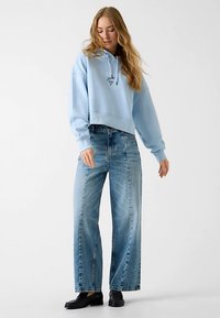 Sweat à capuche court bleu clair avec un logo devant, associé à un jean large délavé. Le mannequin porte des chaussures noires.