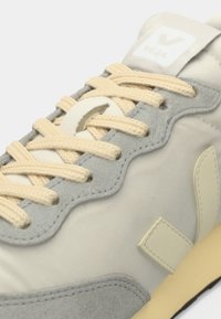 Ανοιχτό γκρι αθλητικό παπούτσι με μπεζ λεπτομέρειες, ανάγλυφο ανώτερο μέρος από σουέτ, κρεμ κορδόνια, επενδεδυμένη γλώσσα και εμφανές λογότυπο V στο πλάι.