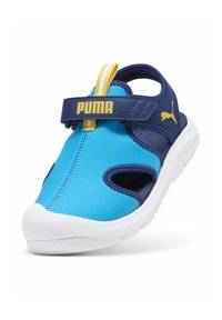 Blå och marinblå sandal med en vit sula, utrustad med en kardborreförslutning, utskärningsdesign och en gul Puma-logotyp på remmen.