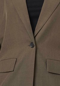 Brun blazer med slagkrave, enkelt sort knaplukning og to forlommer. Stoffet har en glat tekstur med en skræddersyet pasform.