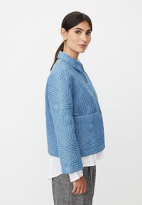 Blå quiltet jakke med geometrisk mønster, med krave, forreste knapper og tekstureret stof, båret over en hvid skjorte.