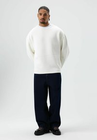 Witte oversized sweater met verlaagde schouders, gecombineerd met donkere wijde jeans en zwarte veterschoenen, staand tegen een effen achtergrond.