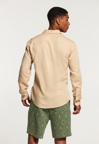 Uomo di spalle che indossa una camicia a maniche lunghe beige e pantaloni corti verdi con un motivo a rombi, su uno sfondo semplice.