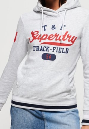 Jasnoszara bluza z kapturem z czerwonym i granatowym napisem "Superdry Track-Field 54", przednią kieszenią, białymi sznurkami i paskowanym dołem, noszona z niebieskimi jeansami.