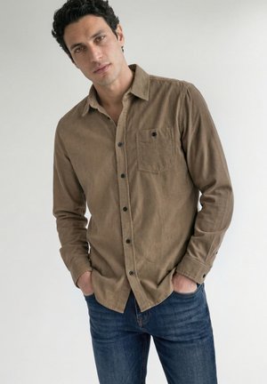Mand med krøllet sort hår iført en brun skjorte med knapper og blå jeans, står med hænderne i lommerne mod en ensfarvet baggrund.