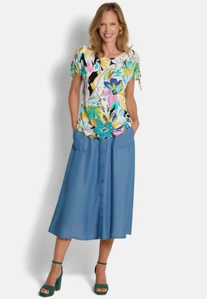 Femme souriante portant un haut coloré à fleurs à manches courtes, une jupe midi bleue boutonnée sur le devant et des sandales vertes à talons hauts, les mains dans les poches.