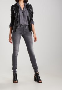 Chaqueta de cuero negra, camisa gris de botonadura, jeans grises ajustados y tacones negros de punta abierta con un tacón ancho, que presentan un detalle de cremallera.