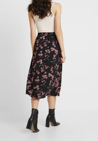 Jupe midi florale en tissu noir ornée de motifs floraux roses et verts, avec une taille ajustée et une silhouette fluide. Associée à des bottes à talons noires.