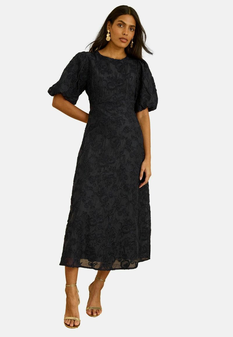 Zwarte jurk van middellange lengte met een gestructureerd bloemenpatroon, pofmouwen en een rechte silhouet. Bijpassende gouden sandalen.