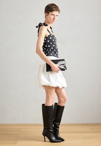 Haut à pois noir et blanc avec liens aux épaules, jupe bouffante blanche, pochette noire et bottes noires à talons aiguilles montantes. Textures lisses.