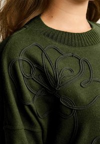 Zielony sweter o teksturowanym splocie z wyraźnym, haftowanym kwiatowym wzorem w dopasowanym kolorze nici. Żebrowany dekolt i rękawy.