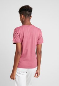 Tričko s krátkým rukávem, kulatým výstřihem v jemně růžové bavlněné látce s ohrnutými rukávy a hladkou texturou. Bez viditelných vzorů nebo kovových prvků.