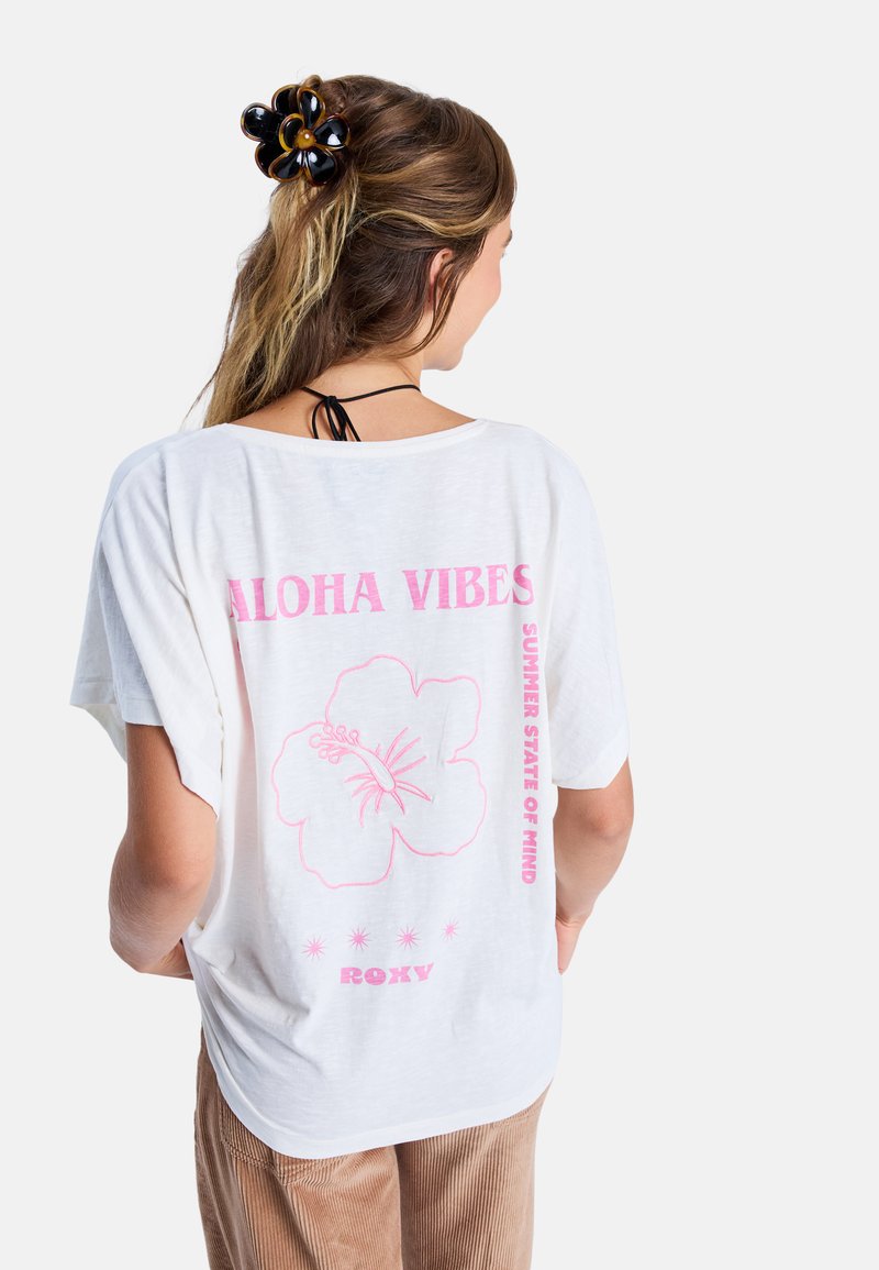 Bílý volný krátký rukáv tričko s růžovou květinovou grafikou a textem "ALOHA VIBES" na zádech, doplněné o texturované hnědé kalhoty.