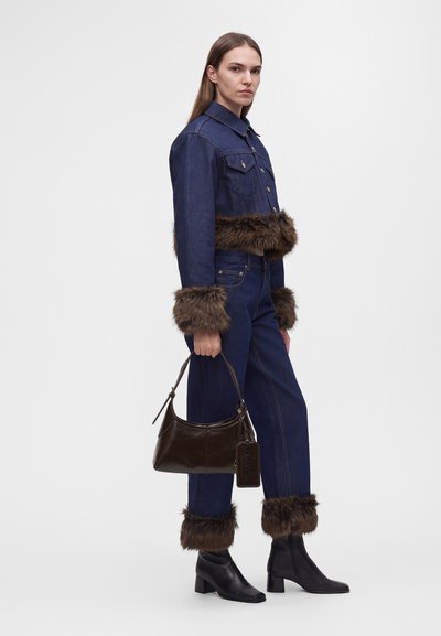 Femme en veste et pantalon en denim bleu foncé avec une bordure en fourrure marron aux poignets et à la taille, tenant un sac à main marron foncé et portant des bottines noires.