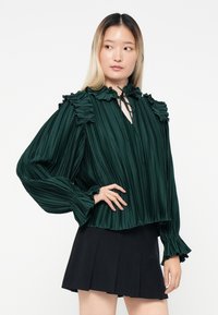 Blusa plissettata verde scuro con dettagli arricciati sulle spalle e sui polsini, dotata di scollatura a chiave e dettaglio annodato. Abbinata a una gonna nera.