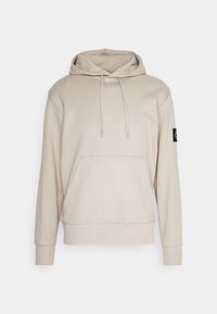 Sudadera beige hecha de una tela suave, con un bolsillo tipo canguro, capucha con cordón, puños acanalados y un parche negro con el logo en la manga.