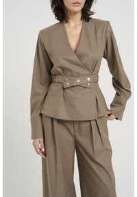 Haut cache-cœur marron à manches longues avec un décolleté en V profond, doté d'une taille ceinturée avec des boutons argentés. Associé à un pantalon large assorti.