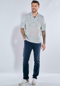 Hellgrau gestreiftes Langarmshirt mit Kapuze, kombiniert mit dunkelblauen Jeans und beigen Sneakers. Zeigt Tattoos an den Armen.