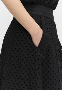 Robe noire en tissu texturé avec des motifs de points ouverts, une taille lisse et des poches latérales pour plus de fonctionnalité.