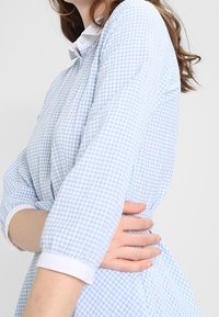 Kvinna i en blå och vit rutad klänning med vit krage och manschetter, som håller sig om midjan med en hand, mot en enfärgad bakgrund.