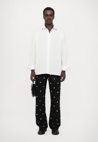 Cămașă albă cu accente roșii, asortată cu pantaloni negri cu modele de puncte argintii. Modelul ține o geantă mică neagră.