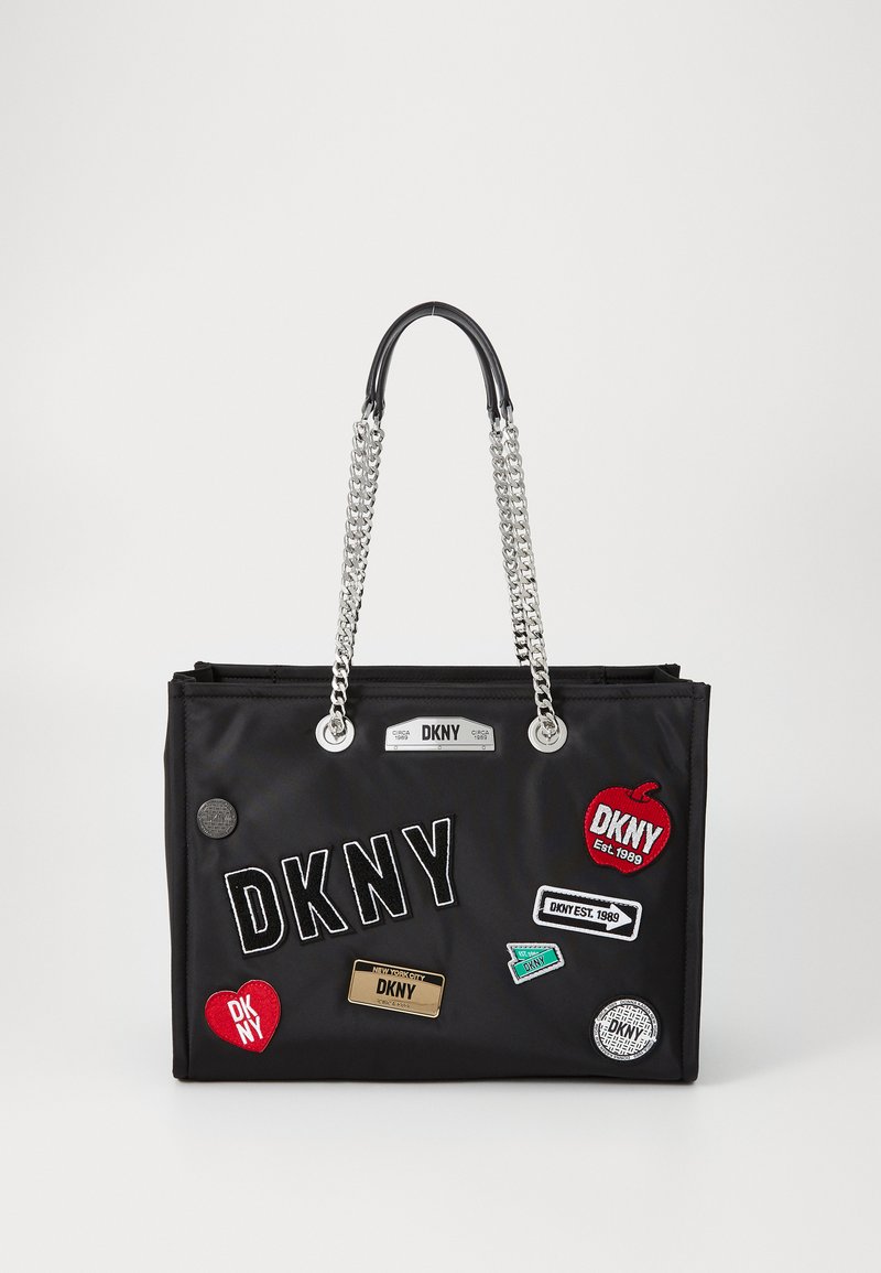 Black nylon tote bag with silver chain handles, featuring multiple colorful patches and a logo plate. Rectangular shape with a zip closure.
