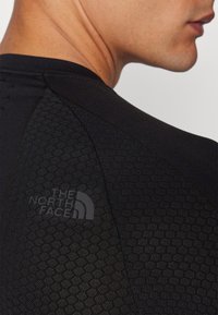 Schwarzes Sportshirt mit strukturiertem Muster, das eine taillierte Passform bietet und ein Logo in Dunkelgrau auf der linken Schulter trägt. Glatte Stoffoberfläche.