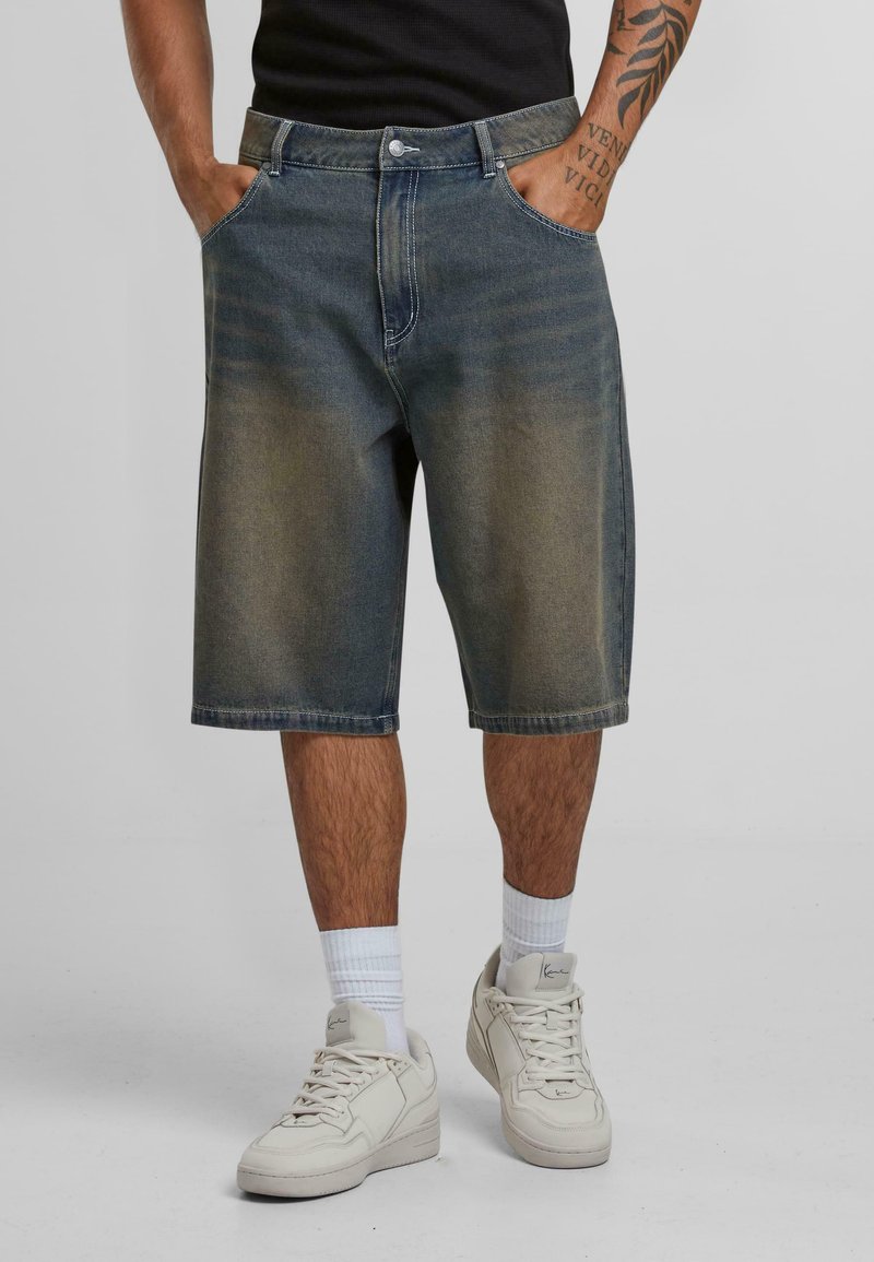 Denim shorts in a faded blue wash, knee-length, with front pockets and a button closure. Paired with white sneakers and ankle-length socks.