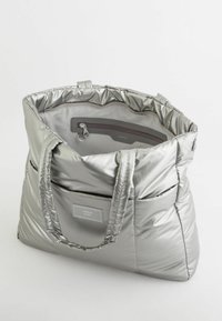 Srebrna torba typu tote z pikowaną teksturą, z dwiema rączkami, wewnętrzną kieszenią na zamek oraz przednią etykietą do brandingu.