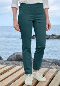 Teal, high-waisted broek gemaakt van een gladde stof, met een getailleerde pasvorm en rechte pijpen. Combinatie met witte casual sneakers op houten vlonder.