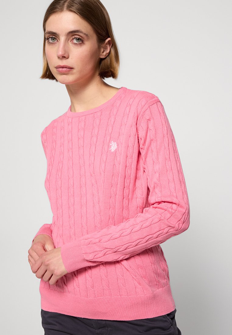 Junge Frau mit kurzen Haaren, die einen rosa Kabelstrickpullover und dunkle Hosen trägt, steht vor einem hellen, einfarbigen Hintergrund.