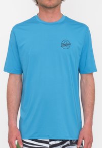 Camisa de manga corta azul claro hecha de una tela ligera y suave. Presenta un logotipo negro circular en el lado izquierdo del pecho. Cuello redondo estándar.