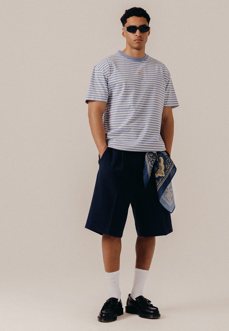 Homme portant des lunettes de soleil noires, un t-shirt rayé bleu et blanc, un short bleu marine avec une écharpe à motifs, des chaussettes blanches et des chaussures noires sur un fond uni.